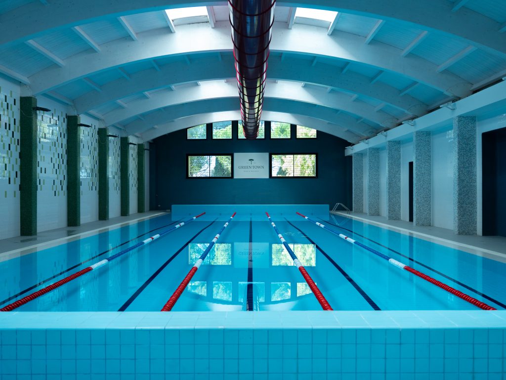 Piscina coperta con acqua limpida e blu, divisori di corsie e un alto soffitto ad arco. Le grandi finestre lasciano entrare la luce naturale all'estremità, mentre l'area della piscina è pulita e vuota.