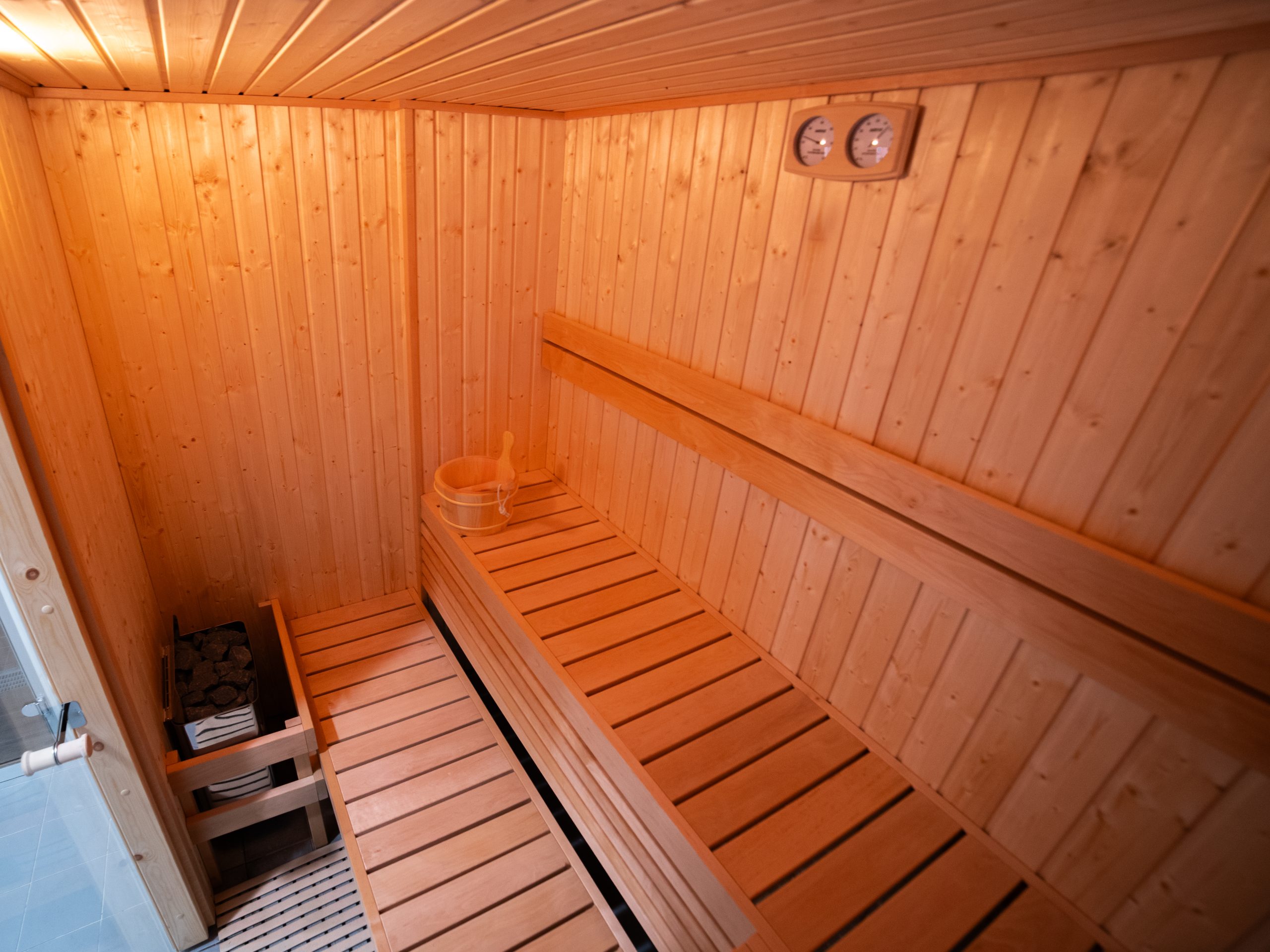 Una sauna in legno con panche lungo le pareti, un secchio con un mestolo sulla panca, una stufa con pietre nell'angolo, un termometro e un igrometro montati sulla parete.