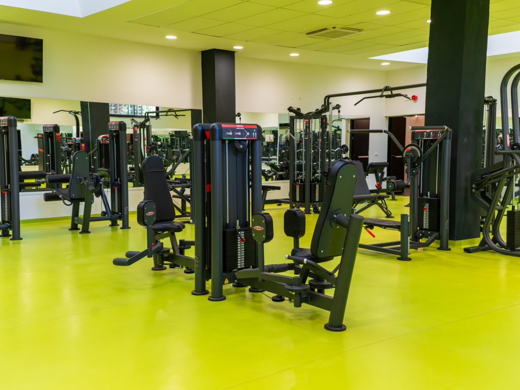 Una palestra moderna con pavimenti gialli, diverse macchine per l'allenamento della forza e grandi specchi alle pareti. Lo spazio è pulito, spazioso e ben illuminato, senza persone.