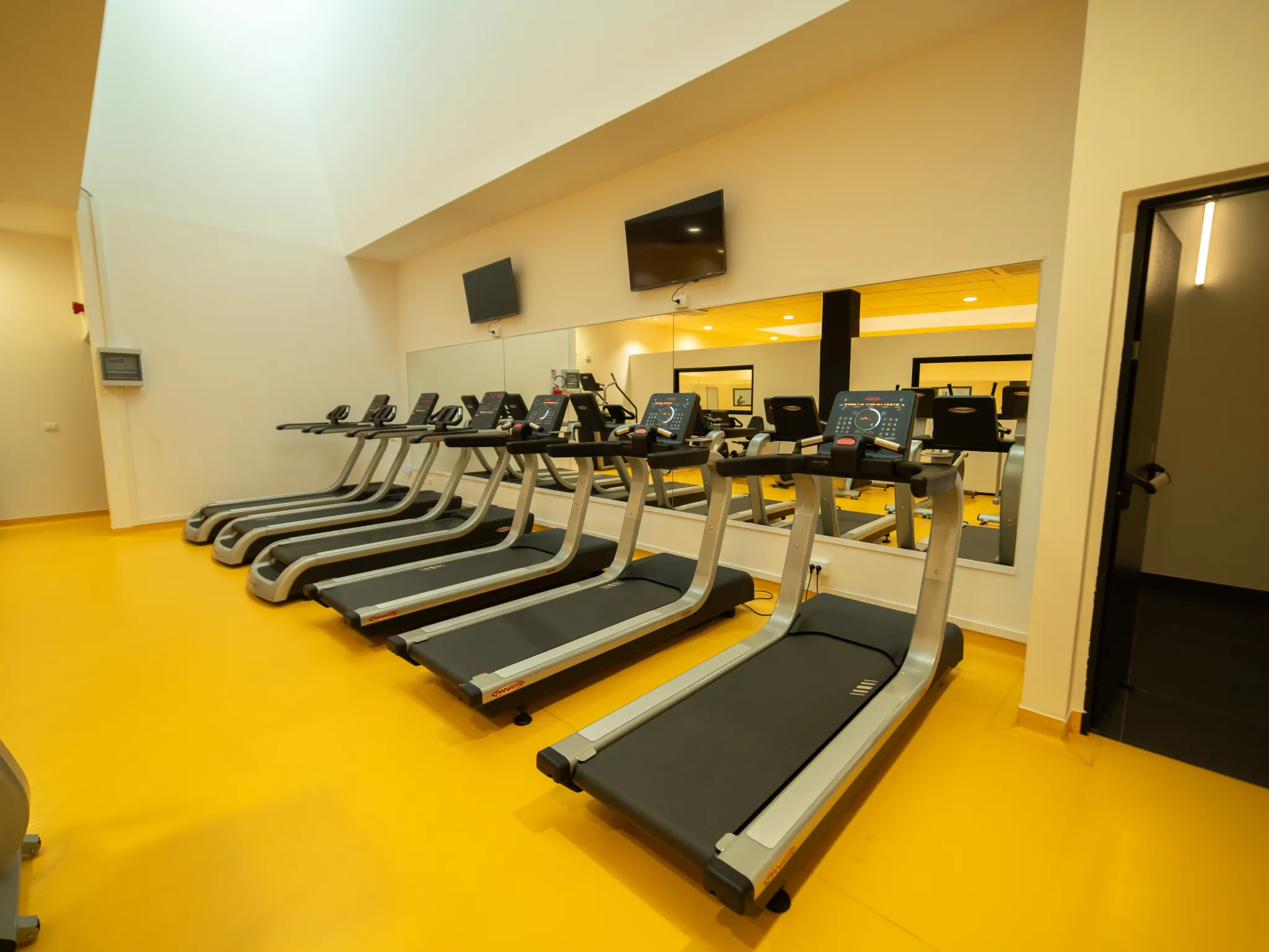 Una fila di tapis roulant è allineata in una palestra con pavimenti giallo brillante, grandi specchi alle pareti e due televisori a muro sopra i tapis. L'ambiente è ben illuminato e moderno.