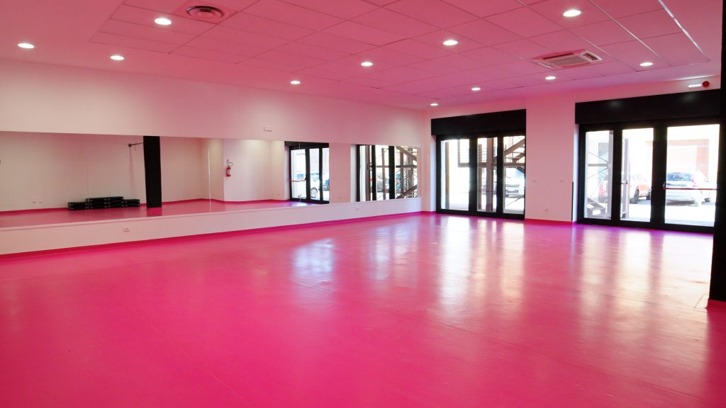 Un'ampia sala da ballo con pavimenti rosa acceso, pareti bianche, grandi specchi a muro, luci a soffitto e finestre a tutta altezza che lasciano entrare la luce naturale dall'esterno.